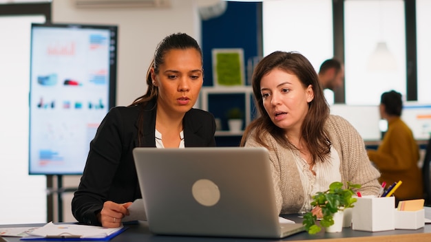 Gerente de proyecto joven y director de la empresa hablando de estrategia empresarial frente a la computadora portátil en la oficina creativa. Equipo diverso de compañeros de trabajo que trabajan en el ajetreado lugar de trabajo de la agencia de marketing.