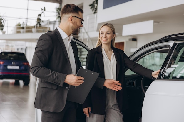 Gerente de ventas de automóviles mostrando el automóvil a una compradora caucásica de pie en un concesionario de automóviles de lujo