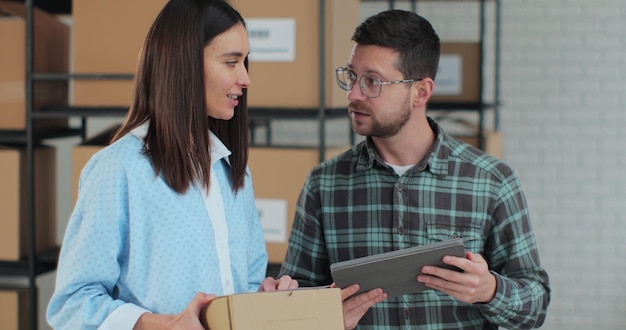 Un gerente de ventas que usa una tableta está hablando con un empleado que sostiene una caja de cartón Un almacén con paquetes listos para su envío en segundo plano