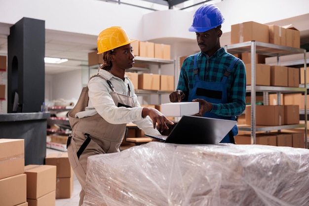 Gerentes de armazéns discutindo a escolha da caixa para o pedido do comprador. Funcionário do serviço de transporte apontando para o laptop e analisando a logística de entrega de mercadorias com o assistente de armazém afro-americano