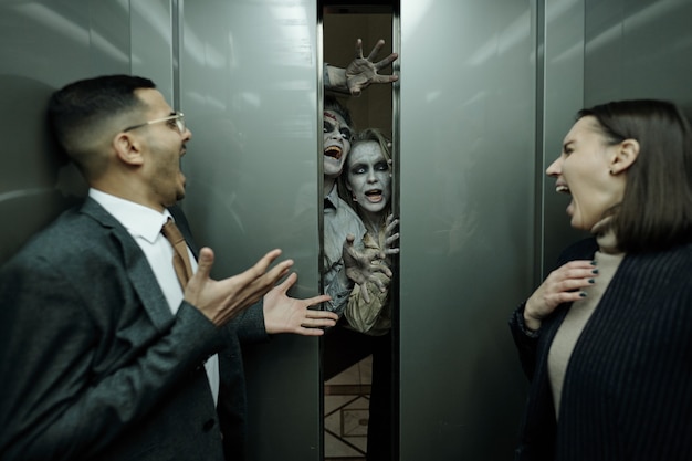 Gerentes de oficina emocionados contemporáneos jóvenes en ropa formal gritando de horror en el ascensor en movimiento mientras dos zombies intentan entrar