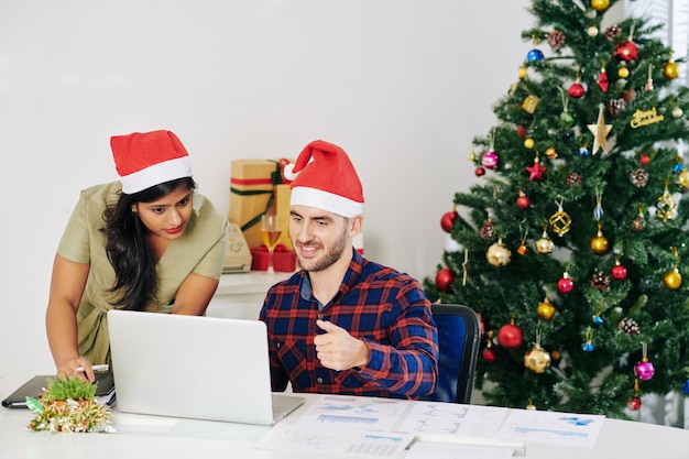 Gerentes de proyecto en sombreros de Santa Claus discutiendo el informe en la pantalla del portátil en la oficina decorada