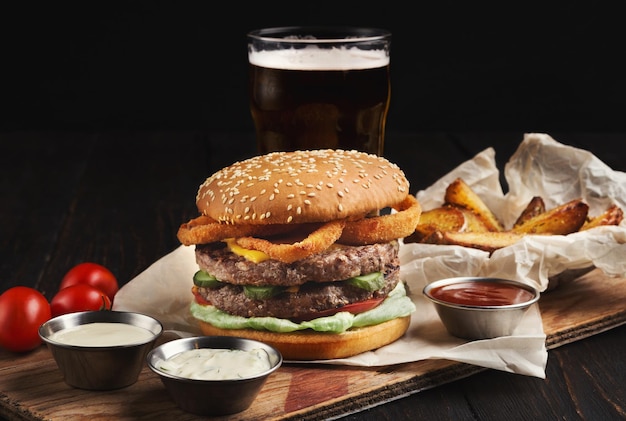 Gericht im Fast-Food-Restaurant. Saftiger Burger, Kartoffelecken, Saucen und ein Glas kaltes Bier auf dunklem Holzhintergrund