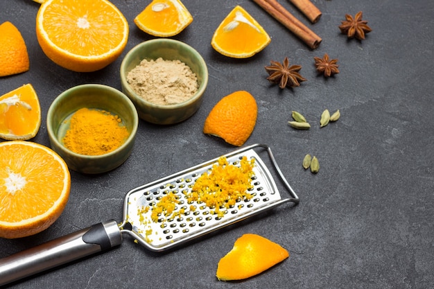 Geriebene Orangenschale auf Reibe. Geschnittene Orange und Gewürze auf dem Tisch. Schwarzer Hintergrund. Ansicht von oben.