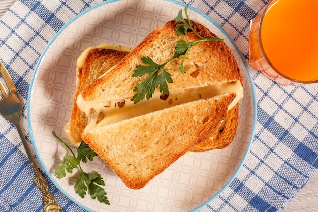 Geröstete Brotscheiben mit Käse und grüner Petersilie auf weißem Teller, Glas Orangensaft mit blauer Küchenserviette. Gutes Essen zum Frühstück. Ansicht von oben