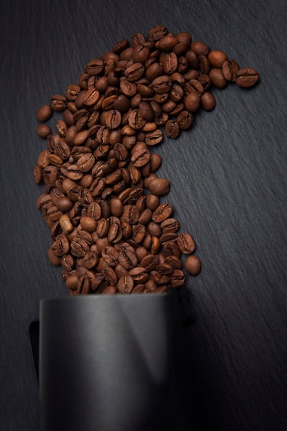 Geröstete Kaffeebohnen, die aus dem schwarzen Milchkännchen verschüttet wurden Zubereitung eines aromatischen Getränks Closeup Vertikal Dunkler Hintergrund