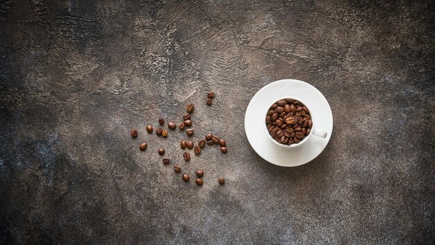 Geröstete Kaffeebohnen in weißer Tasse auf strukturiertem Steinhintergrund mit Kopierraum, Ansicht von oben