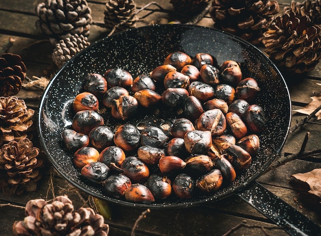Geröstete kastanien in einer kastanienpfanne mit löchern auf einem tisch mit herbstblättern und tannenzapfen