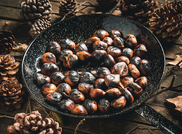 Geröstete Kastanien in einer Kastanienpfanne mit Löchern auf einem Tisch mit Herbstblättern und Tannenzapfen