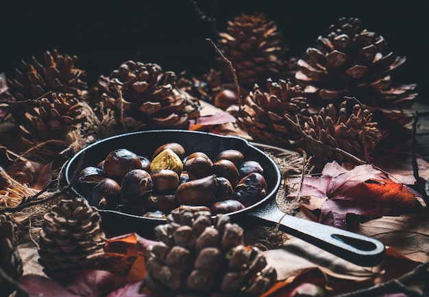 Geröstete Kastanien in einer kleinen Eisenpfanne auf einem Tisch mit Herbstlaub und Tannenzapfen