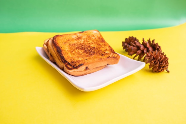 Geröstetes Brot Auf Einem Weißen Teller Mit Fichtenblumen Auf Gelbem Und Grünem Papierhintergrund. Toast zum Frühstück. Horizontales Foto