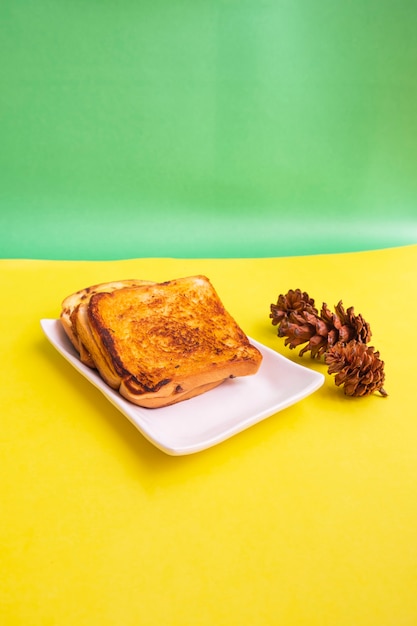 Geröstetes Brot Auf Einem Weißen Teller Mit Fichtenblumen Auf Gelbem Und Grünem Papierhintergrund. Toast zum Frühstück. Vertikales Foto