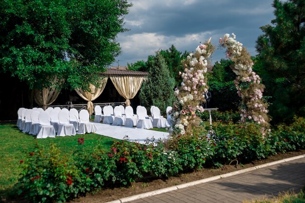 Foto gesamtansicht der dekoration der hochzeitszeremonie im garten in der luft. hochzeitsdekor ein bogen aus frischen blumen, stühle in decken für die gäste