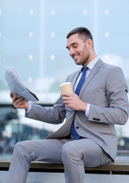 Geschäft, heiße Getränke und Leute und Konzept - junger lächelnder Geschäftsmann mit Papierkaffeetasse und -zeitung über Bürogebäude