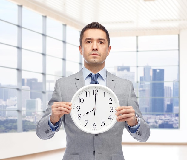 Geschäft, Leute, Zeitmanagement und Bürokonzept - Geschäftsmann im Anzug, der die Uhr hält, die 8 Uhr oben über dem Fensterhintergrund des Stadtbüros zeigt