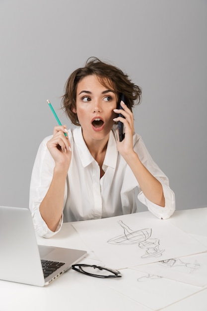 Geschäft schockierte Frau, die lokalisiert über graue Wand beim Sitzen am Tisch unter Verwendung der Laptop-Zeichnung, die per Telefon spricht, posiert
