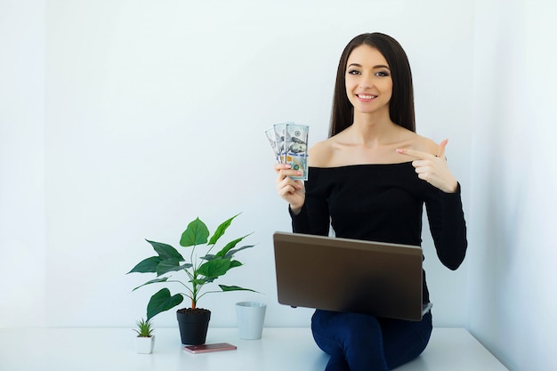 Geschäft. Schöne Geschäftsfrau, die in den Geld-Händen hält. Sitzt auf dem Tisch im Big Light Office und arbeitet am Computer. Happy Girl arbeitet zu Hause. Hohe Auflösung