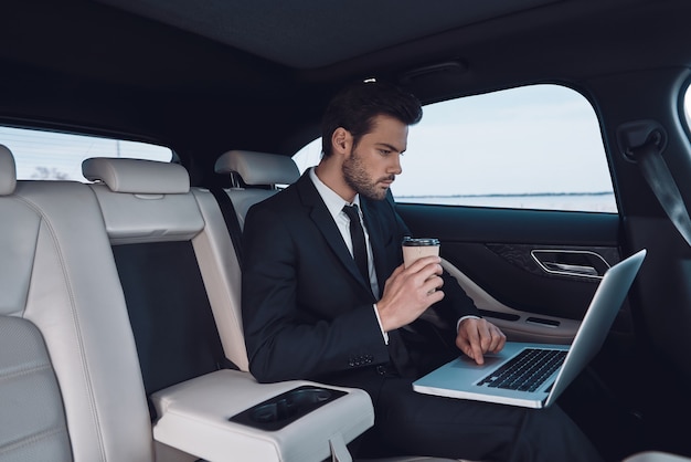 Geschäft unterwegs. Hübscher junger Mann im vollen Anzug, der mit Laptop arbeitet, während er im Auto sitzt