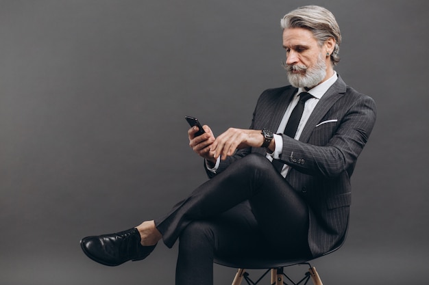 Geschäftlicher und modischer bärtiger reifer Mann in einem grauen Anzug, der Telefon an der grauen Wand sitzt und benutzt.