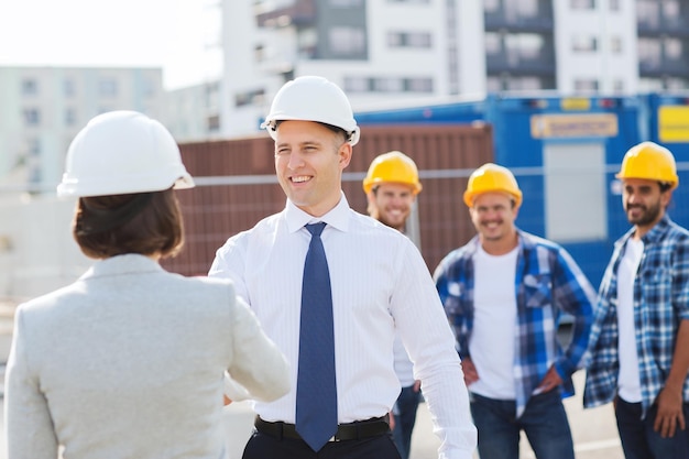 Geschäfts-, Gebäude-, Teamwork-, Gesten- und Personenkonzept - eine Gruppe lächelnder Bauarbeiter in Hardhats mit Klemmbrett, die sich im Freien begrüßen