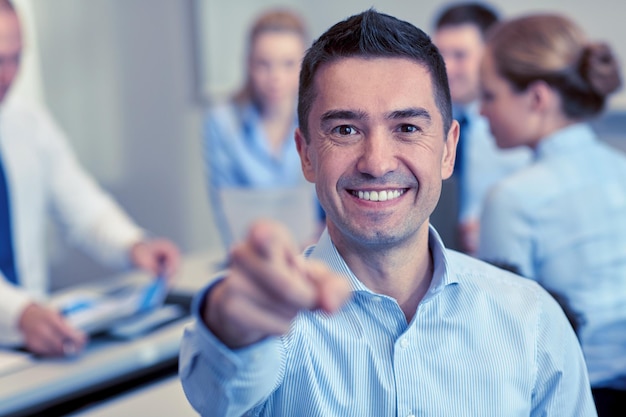 Geschäfts-, Menschen-, Gesten- und Teamwork-Konzept - lächelnder Geschäftsmann, der mit einer Gruppe von Geschäftsleuten, die sich im Büro treffen, mit dem Finger auf Sie zeigt