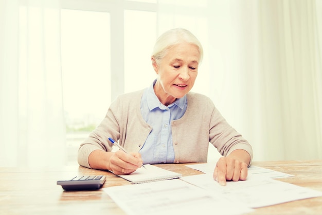 Geschäfts-, Spar-, Rentenversicherungs-, Alters- und Personenkonzept - Seniorin mit Papieren oder Rechnungen und Taschenrechner, die zu Hause schreiben