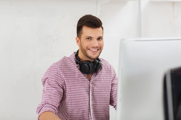 Geschäfts-, Startup-, Bildungs-, Technologie- und Personenkonzept - glücklicher kreativer Mann oder Student mit Kopfhörern und Computer im Büro