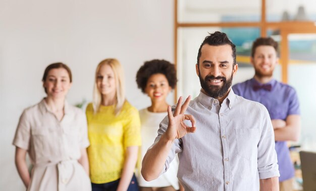 Geschäfts-, Startup-, Menschen-, Gesten- und Teamwork-Konzept - glücklicher junger Mann mit Bart, der ein ok-Handzeichen über dem Kreativteam im Büro zeigt