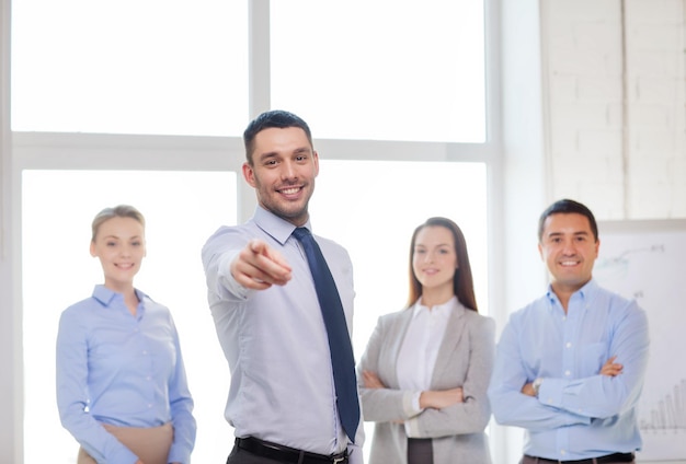 Geschäfts- und Bürokonzept - lächelnder gutaussehender Geschäftsmann mit Team im Büro, das mit dem Finger auf Sie zeigt