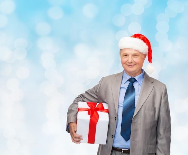 Geschäfts-, Weihnachts-, Geschenk- und Personenkonzept - lächelnder älterer Mann im Anzug und Weihnachtsmann-Helfermütze mit Geschenk über Blaulichthintergrund