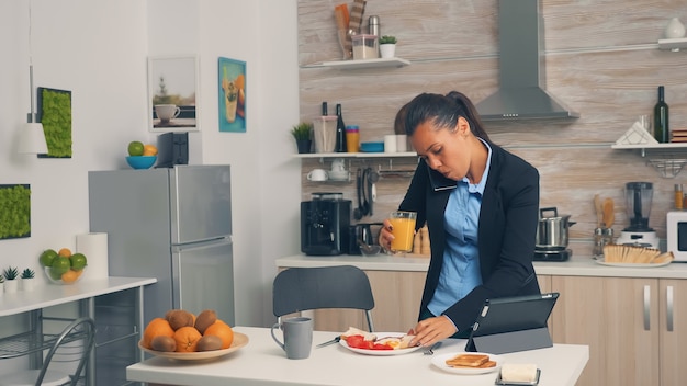 Geschäftsfrau am Telefon beim Frühstücken. Konzentrierte Geschäftsfrau morgens Multitasking in der Küche bevor es ins Büro geht, stressige Lebensweise, Karriere und Ziele zu erreichen