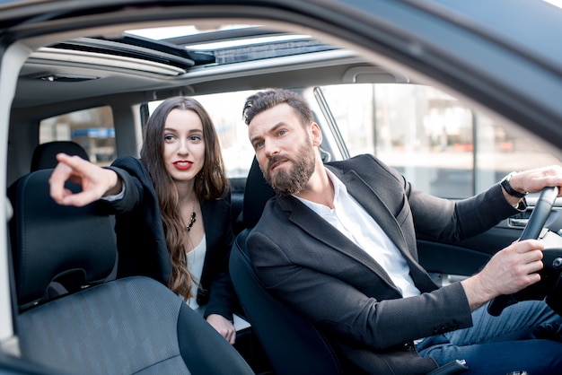 Geschäftsfrau auf dem Rücksitz mit elegantem Mann, der ein Auto in der Stadt fährt. Besorgt über eine Verzögerung bei einem wichtigen Meeting