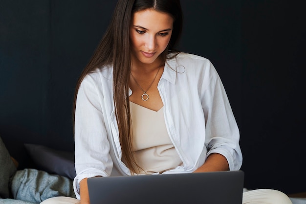 Foto geschäftsfrau benutzt laptop, arbeitet fern von zu hause aus. das lächelnde mädchen im weißen hemd sitzt auf dem bett neben dem laptop. schöne brünette auf dunkler wand der wand im schlafzimmer.