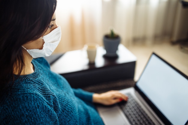 Geschäftsfrau bleibt zu Hause und arbeitet während der Quarantäne der Coronavirus-Epidemie. Arbeiterin, die eine medizinische Maske trägt und auf einem Laptop tippt. Konzept zur Verhinderung der Verbreitung von Covid-19-Pandemien.