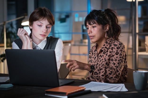 Geschäftsfrau, die auf den Bildschirm des Laptops zeigt und mit ihrem Kollegen über die Online-Präsentation diskutiert, arbeitet sie bis zum späten Abend im Büro