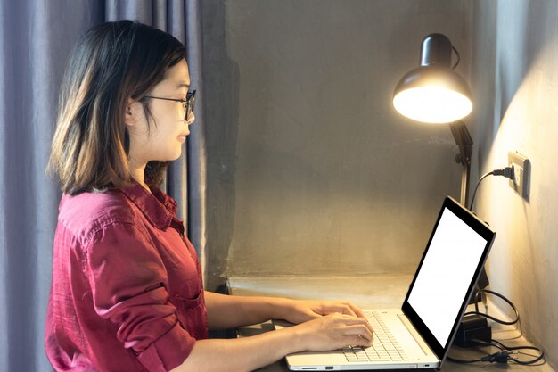 Foto geschäftsfrau, die auf laptop mit leerem weißem schirm für spott herauf schablonenhintergrund mit lampe schreibt