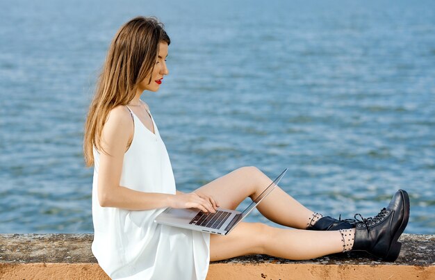 Geschäftsfrau, die bequem auf einem Betonzaun-Anlegeplatz positioniert ist, arbeitet hinter einem Laptop. Foto in hoher Qualität