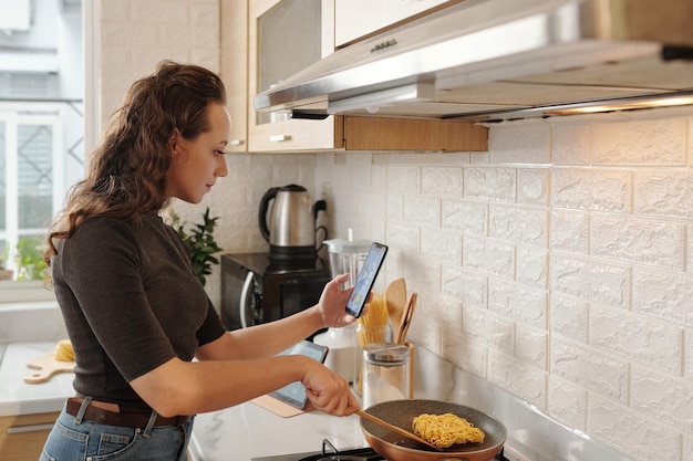 Geschäftsfrau, die den Verkaufsbericht auf dem Smartphone-Bildschirm analysiert, wenn sie das Mittagessen am Küchenherd kocht