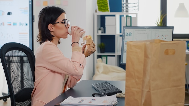 Geschäftsfrau, die einen Bissen eines leckeren Sandwiches isst und Kaffee vor dem Monitor trinkt