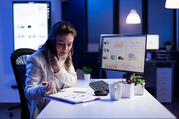 Geschäftsfrau, die Finanzdiagramme überprüft, während sie spät nachts im Firmenbüro mit ihrem Team auf dem Smartphone spricht