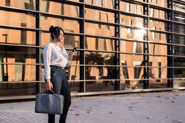 Geschäftsfrau, die ihr Smartphone vor einem Bürogebäude im Finanzviertel konsultiert, Konzept des erfolgreichen Unternehmers und des urbanen Lebensstils, Platz für Text kopieren