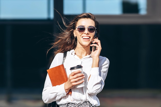 Geschäftsfrau, die im Freien in der Nähe des modernen Business Centers mit Notebook auf dem Handy spaziert und spricht