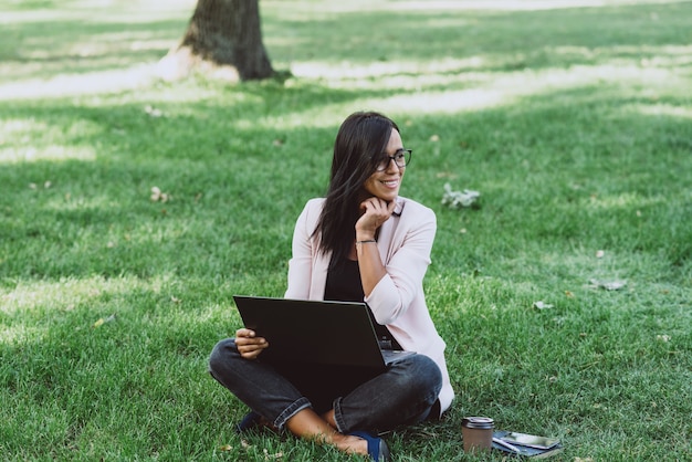 Geschäftsfrau, die im Sommergraspark unter Verwendung des Laptops sitzt.