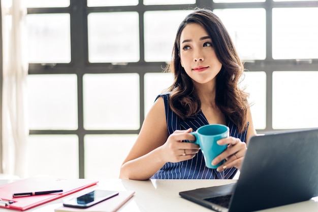 Geschäftsfrau, die mit Laptop-Computer sitzt und arbeitet und Kaffee trinkt. Kreative Geschäftsleute, die in ihrer Arbeitsstation am modernen Arbeitsloft planen