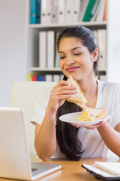 Geschäftsfrau, die Sandwich beim Betrachten des Laptops hat