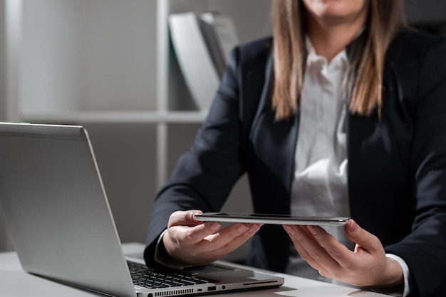 Geschäftsfrau, die Tablet in einer Hand hält und mit anderen auf dem Laptop tippt Sitzende Frau im Büro, die Informationen auf dem Tablet präsentiert und auf dem Computer schreibt