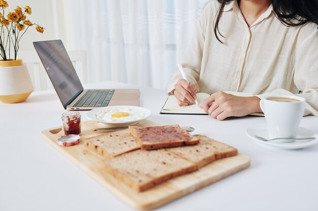 Geschäftsfrau, die zu Hause frühstückt und im Planer schreibt, bevor sie sich mit Kollegen online trifft