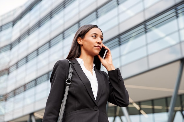 Geschäftsfrau, die zur Arbeit pendelt und am Handy außerhalb des modernen Bürogebäudes spricht