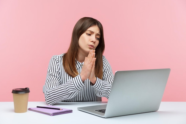 Geschäftsfrau im gestreiften Hemd, die Hände in betender Geste hält und über einen Videoanruf auf dem Laptop spricht, herzlich fragt, mit verzweifeltem Gesicht fleht. Studioaufnahme im Innenbereich isoliert auf rosa Hintergrund