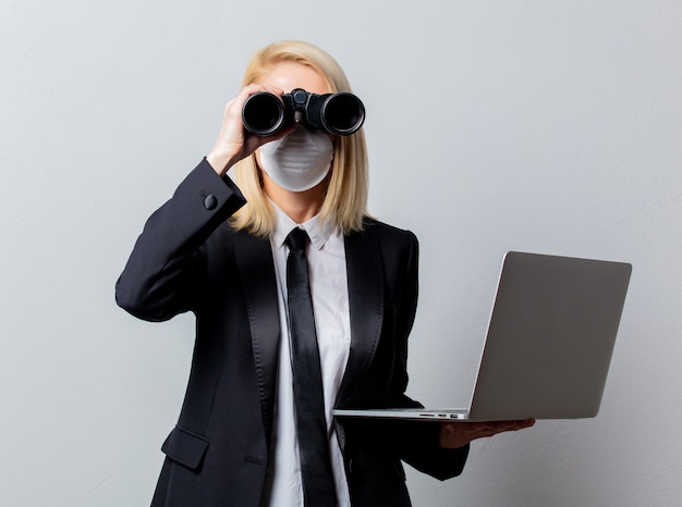 Geschäftsfrau im schwarzen Anzug und in der Gesichtsmaske mit Fernglas und Computer